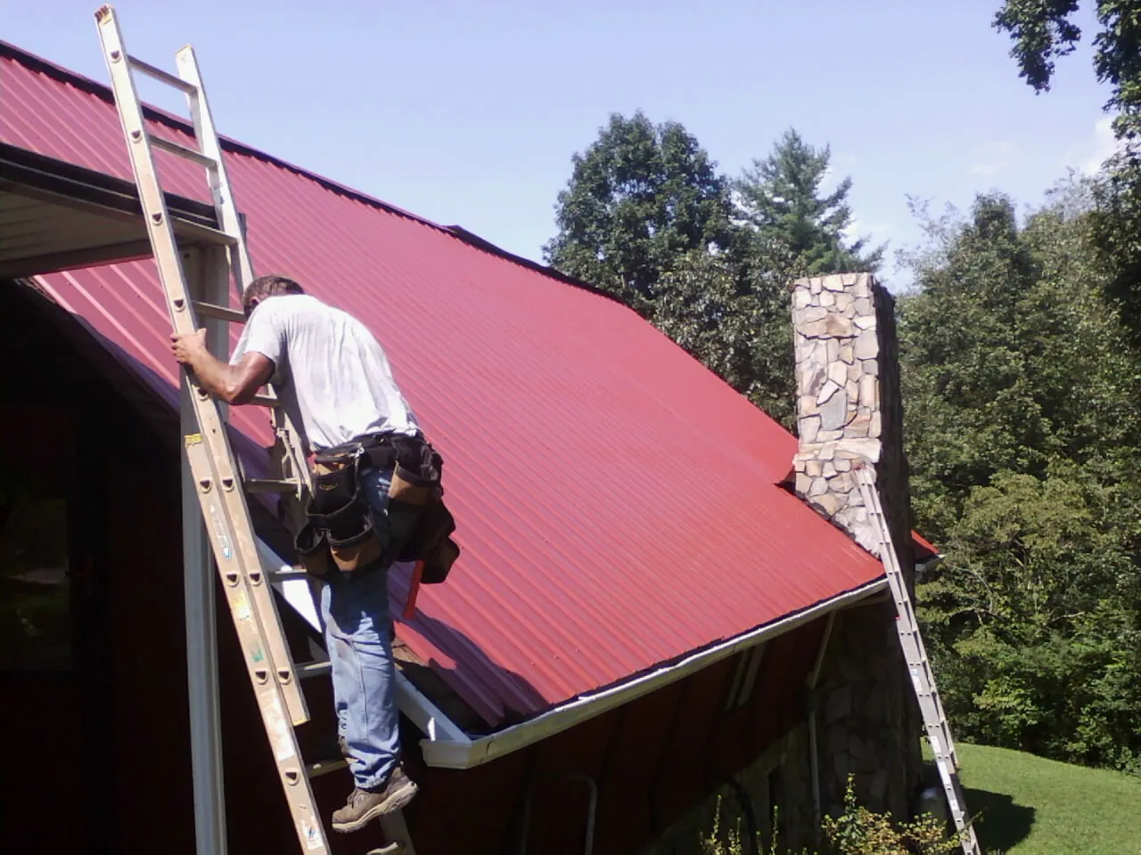 Roofer on a ladder beside a red metal roof and stone chimney — Soffit & Fascia Repair by Roofing Brothers in Murrells Inlet